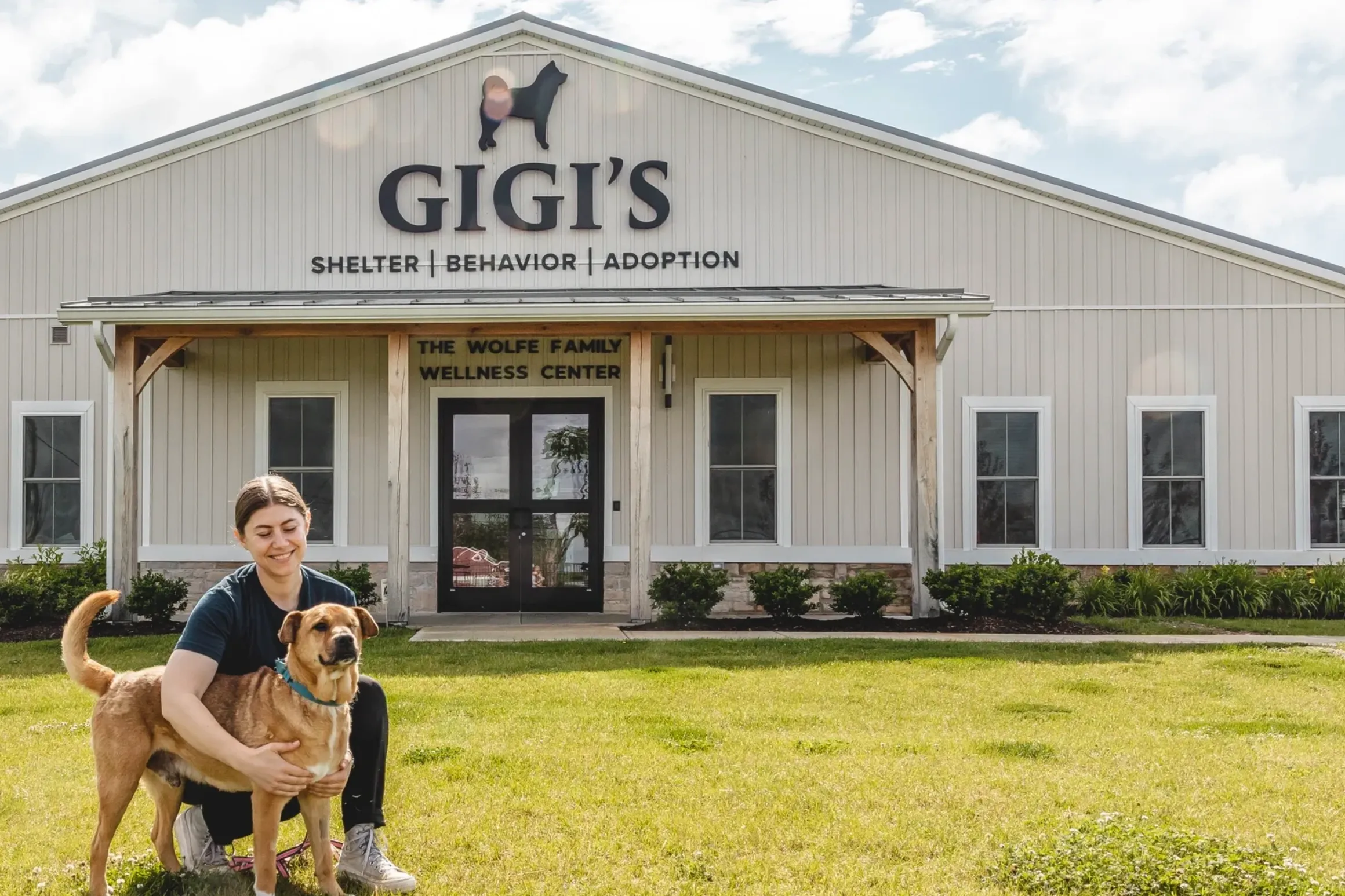 Exterior of Gigi's animal shelter and adoption center, featuring a barn-style building with “GIGI'S Shelter | Behavior | Adoption” signage. In front of the building, a person kneels on the lawn beside a tan dog, with the shelter entrance and landscaped grounds visible behind them.